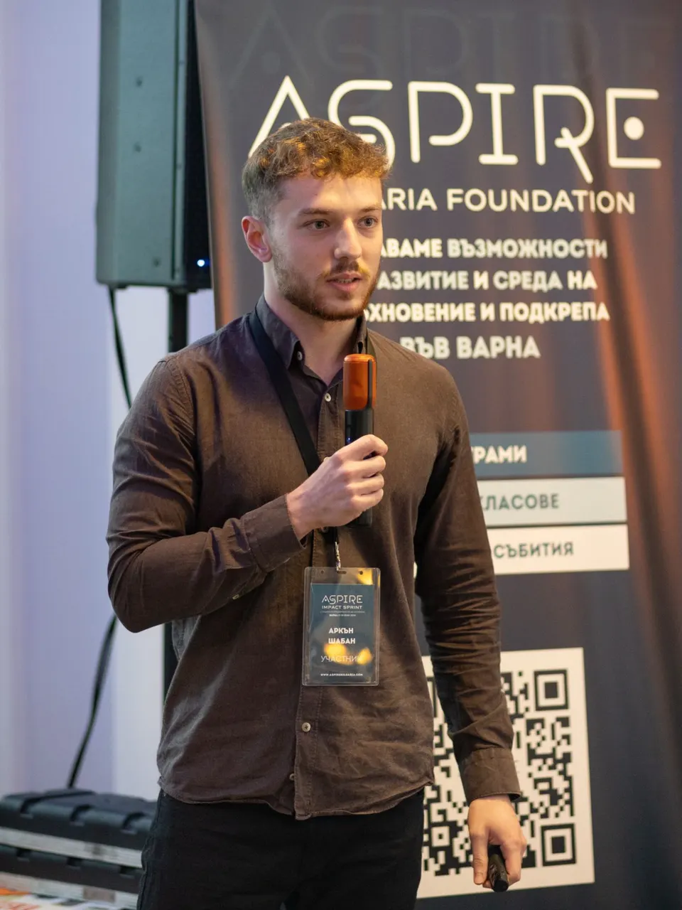 Arkan pitching with a microphone in front of the Aspire Bulgaria Foundation banner